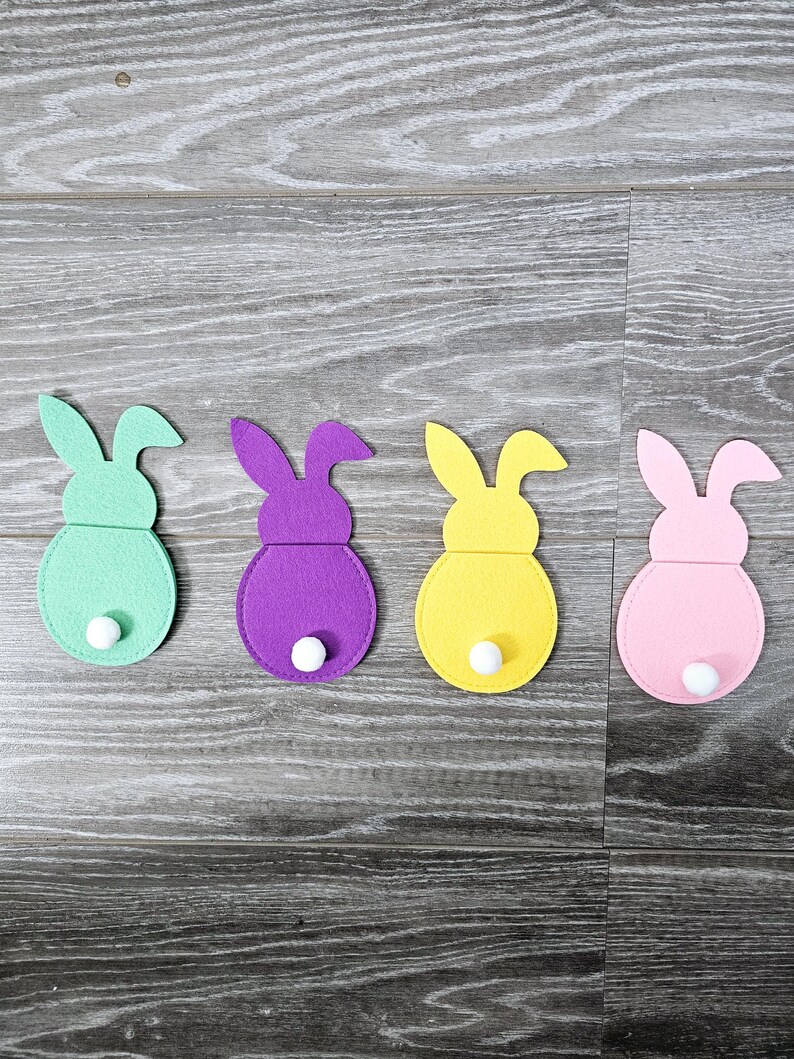 May include: Four felt bunny shapes in different colors: green, purple, yellow, and pink. Each bunny has a white pom-pom tail.