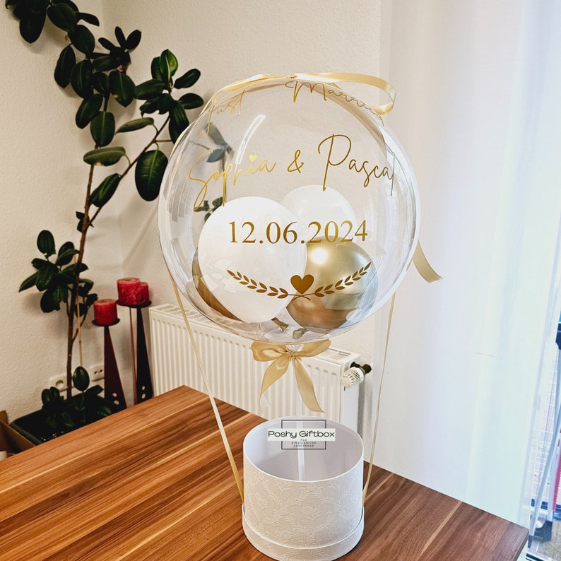 May include: A clear balloon hot air balloon with gold and white balloons inside. The balloon is decorated with a gold ribbon and has the text "Sophia & Pascal 12.06.2024" written on it. The balloon is sitting on top of a white gift box with the text "Poshy Giftbox" on it.