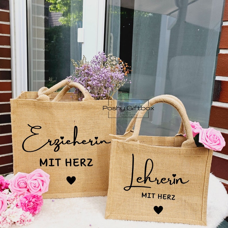 May include: Two brown burlap tote bags with black text. The larger bag says "Erzieherin mit Herz" with a small black heart. The smaller bag says "Lehrerin mit Herz" with a small black heart. Both bags are sitting on a wooden surface with pink flowers.
