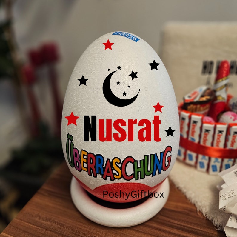 May include: A white egg-shaped container with a red star, black crescent moon, and black stars. The container has the word "Nusrat" in red letters and the word "Ueberraschung" in rainbow letters. The container is sitting on a white base with the words "PoshyGiftbox" printed on it.