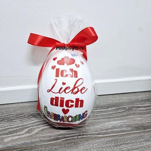 May include: A white chocolate egg wrapped in clear plastic with a red ribbon. The egg has a red heart and the text "Ich Liebe Dich" with a red heart and the text "&Uuml;berraschung" in rainbow colors.