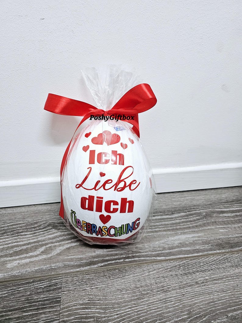 May include: A white chocolate egg wrapped in clear plastic with a red ribbon. The egg has a red heart and the text "Ich Liebe Dich" with a red heart and the text "&Uuml;berraschung" in rainbow colors.