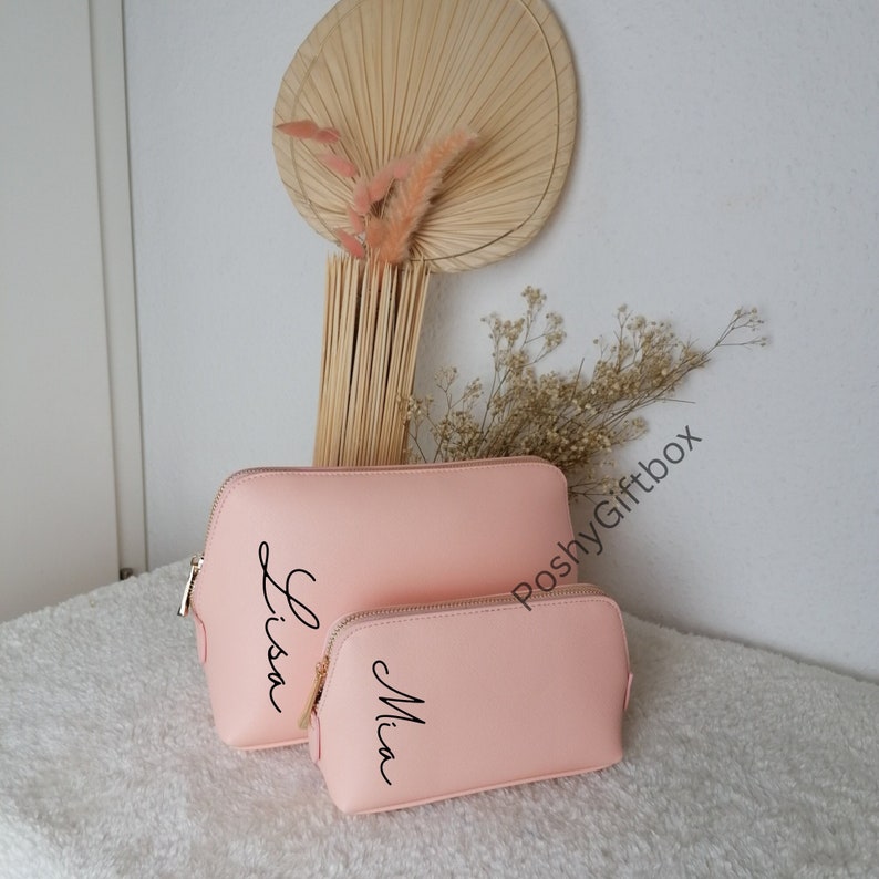 May include: Two pink cosmetic bags with gold zippers. The larger bag has the name "Lisa" written on it in black cursive. The smaller bag has the name "Mia" written on it in black cursive.