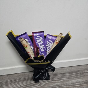 May include: A bouquet of chocolate bars wrapped in black paper with a gold ribbon. The chocolate bars are Milka brand and feature a purple wrapper with a white logo. The bouquet is tied with a black ribbon and has a gold tag that says "PoshyGiftbox".