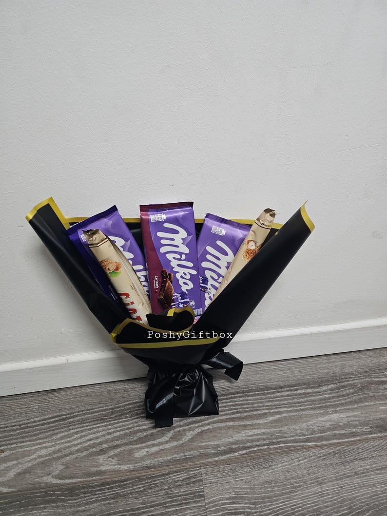 May include: A bouquet of chocolate bars wrapped in black paper with a gold ribbon. The chocolate bars are Milka brand and feature a purple wrapper with a white logo. The bouquet is tied with a black ribbon and has a gold tag that says "PoshyGiftbox".