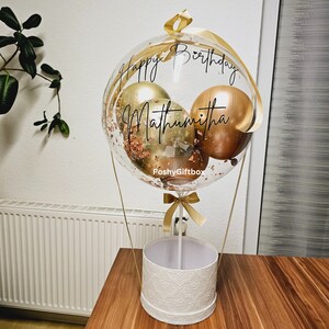 May include: A clear balloon with gold and copper balloons inside, confetti, and the text "Happy Birthday Mathumitha" on a white box with a gold ribbon.