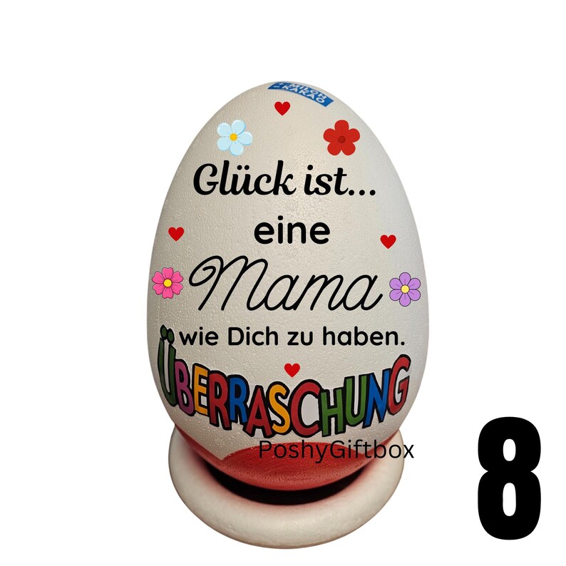 May include: A decorative egg-shaped ornament with the text "Gl&uuml;ck ist... eine Mama wie Dich zu haben. &Uuml;BERRASCHUNG" in black script. The egg is white with red hearts and colorful flower accents. It sits on a white and red base.