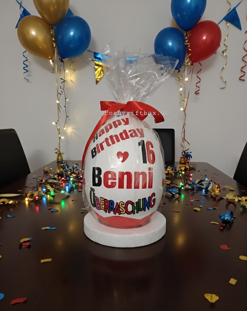 Może przedstawiać: Biały, owalny pakunek prezentowy z napisem "Happy Birthday 16 Benni &Uuml;berraschung" w kolorze czerwonym i czarnym. Pakunek stoi na białym stojaku, otoczony konfetti i balonami w kolorze złotym, niebieskim i czerwonym.