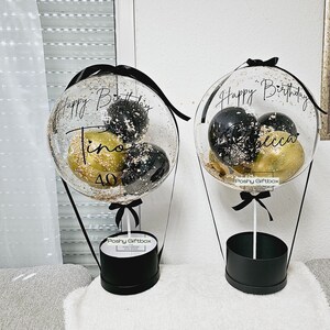 May include: Two clear balloon hot air balloons with black and gold confetti inside. The balloons are tied to black boxes with black ribbon and the text "Poshy Giftbox" printed on them. The balloons have the text "Happy Birthday" and a name on them.