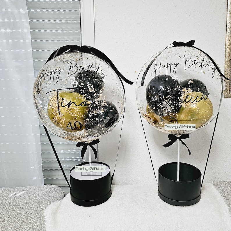 May include: Two clear balloon hot air balloons with black and gold confetti inside. The balloons are tied to black boxes with black ribbon and the text "Poshy Giftbox" printed on them. The balloons have the text "Happy Birthday" and a name on them.