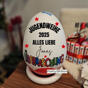 May include: A large white egg-shaped gift box with the text "JUGENDWEIHE 2025 ALLES LIEBE Jonas &Uuml;BERRASCHUNG" in black and colorful lettering. Red star accents are scattered around the text. The egg sits on a white base with a red rim.