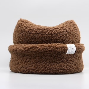 May include: A brown sherpa fleece beanie with a cuffed brim and two pointed cat-ear shapes at the top. A small white rectangular fabric tag is sewn onto the side of the cuff, featuring black line art illustrations of mushrooms.