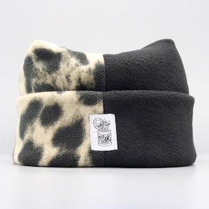 May include: A fleece beanie featuring a split design with one side in solid black and the other in a cream and black animal print. A white rectangular fabric label is sewn on the front, displaying a line drawing of mushrooms in a can.