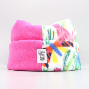 May include: A fleece beanie hat with a split design. One half is bright pink, the other features a colorful floral pattern with pink, blue, yellow, and green on a white background. A small tag is sewn on the side.