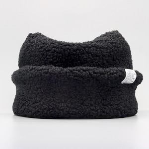 May include: A black beanie made of textured sherpa fleece material. The hat features a rolled brim and two pointed ears at the top. A small white rectangular fabric tag with black text is sewn onto the side of the brim.