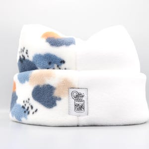 May include: A white fleece beanie hat with a folded brim. The upper section has a pattern of blue, tan, and orange shapes with black accents. A small tag with text is sewn on the front. The hat is designed to keep the head warm.