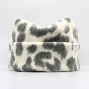 May include: A soft, fleece beanie hat with a gray and white animal print. The hat has a folded brim and a rounded top. A small, silver tag is sewn on the brim. The hat is designed to keep the head warm.