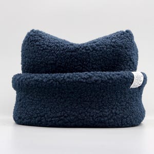 May include: A dark navy blue beanie made of textured sherpa fleece fabric. The hat features a folded cuff and two pointed ears at the top. A small white rectangular fabric tag with black text is attached to the side of the cuff.