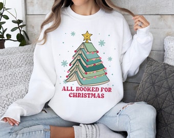 以下が含まれることがあります： 本で作られたクリスマスツリーのデザインが施された白いスウェットシャツ。ピンクとグリーンのオーナメントと金の星で飾られています。「ALL BOOKED FOR CHRISTMAS」の文字がツリーの下にプリントされています。