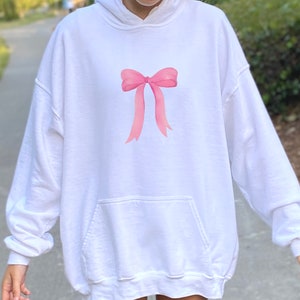 May include: White oversized hoodie with a pink bow graphic on the front. The hoodie has a kangaroo pocket and a drawstring hood.