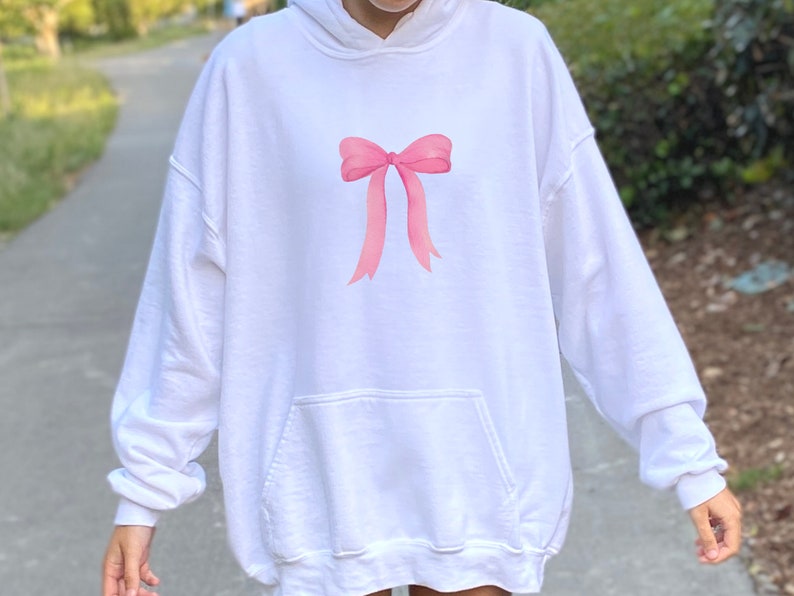 May include: White oversized hoodie with a pink bow graphic on the front. The hoodie has a kangaroo pocket and a drawstring hood.