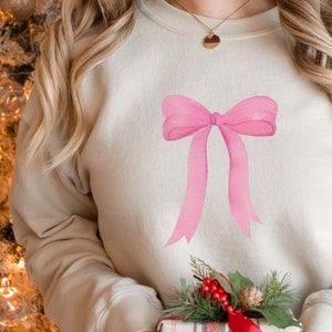 May include: A light beige sweatshirt with a large pink bow design on the front. The sweatshirt is worn by a person with long blonde hair.