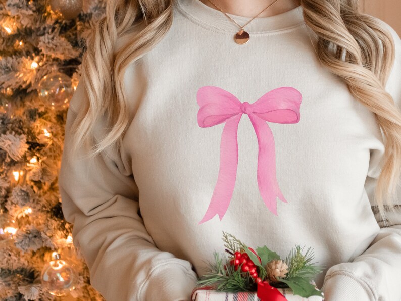 May include: A light beige sweatshirt with a large pink bow design on the front. The sweatshirt is worn by a person with long blonde hair.