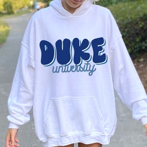 May include: White hoodie with a blue graphic that says "DUKE university" in a retro font.