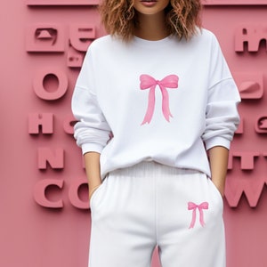 May include: White sweatshirt with a pink bow design on the front and a smaller pink bow design on the left pocket. The sweatshirt is paired with white sweatpants.