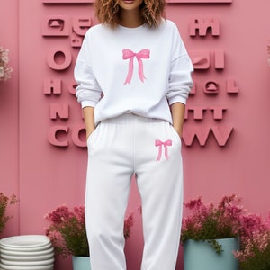 May include: A white sweatshirt and matching sweatpants with pink bow designs. The sweatshirt has a large bow on the front and the sweatpants have a smaller bow on the left leg.