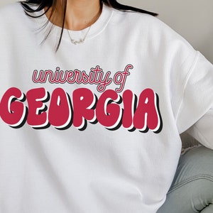 May include: White sweatshirt with a red and black graphic that says "university of GEORGIA" in a retro font.