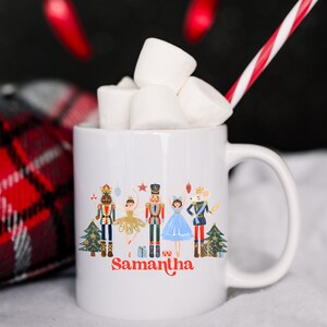 May include: White ceramic mug featuring a colorful illustration of Nutcracker characters, a Christmas tree, and the name "Samantha" in red script. The mug is filled with marshmallows and a candy cane. A red and black plaid blanket is in the background.