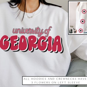 May include: White sweatshirt with the text "university of GEORGIA" in red and black lettering. The sweatshirt has three red flowers on the left sleeve.