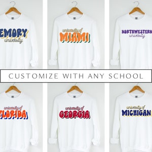 May include: Six white long-sleeved sweatshirts with different university names printed on the front in colorful, retro-style lettering. The text on the sweatshirts reads: "EMORY university", "university of MIAMI", "NORTHWESTERN university", "university of FLORIDA", "university of GEORGIA", and "university of MICHIGAN".