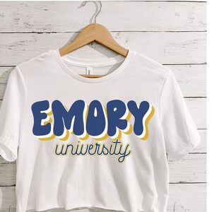 May include: White crop top with short sleeves featuring a blue and yellow graphic that says "EMORY university" in a retro font.