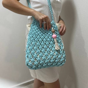 May include: A light blue crocheted tote bag with a pink and brown beaded tassel. The bag is worn over the shoulder of a person wearing a white sleeveless dress.