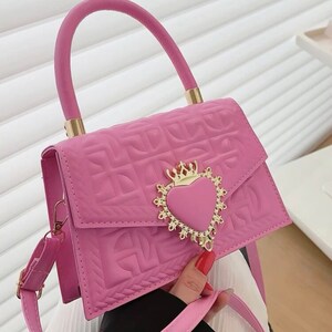 Puede incluir: Un bolso de mano acolchado de color rosa con un cierre dorado en forma de coraz&oacute;n y una corona en la parte superior. El bolso tiene un asa superior y una correa larga para el hombro.