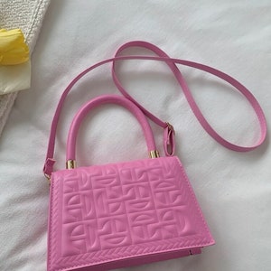 Puede incluir: Un bolso de mano acolchado de color rosa con una correa de cadena dorada y un asa superior. El bolso tiene un patr&oacute;n geom&eacute;trico en relieve en la parte delantera.