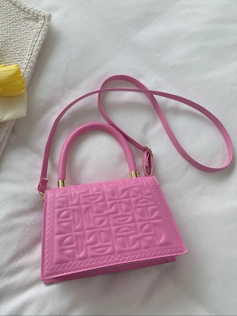 Puede incluir: Un bolso de mano acolchado de color rosa con una correa de cadena dorada y un asa superior. El bolso tiene un patr&oacute;n geom&eacute;trico en relieve en la parte delantera.