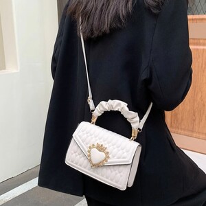 Puede incluir: Bolso blanco acolchado con un cierre dorado en forma de coraz&oacute;n y una correa de cadena dorada. El bolso tiene un asa superior cubierta con tela blanca.