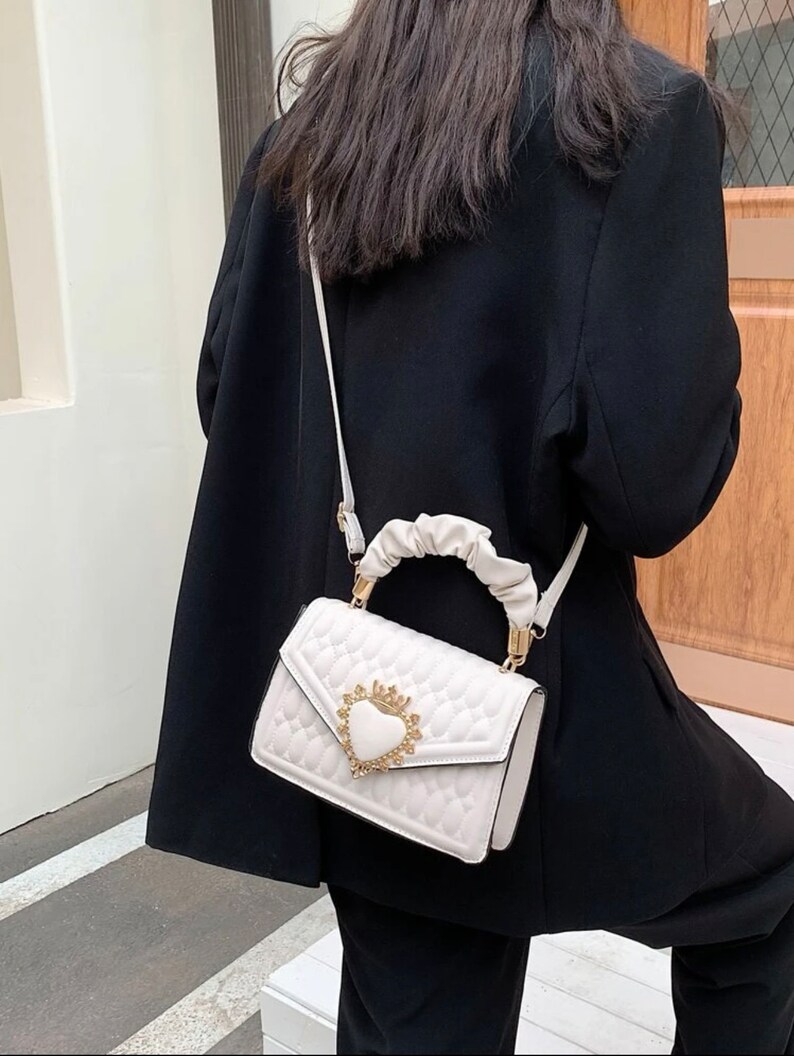 Puede incluir: Bolso blanco acolchado con un cierre dorado en forma de coraz&oacute;n y una correa de cadena dorada. El bolso tiene un asa superior cubierta con tela blanca.