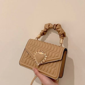 Puede incluir: Un bolso de mano marr&oacute;n acolchado con un cierre dorado en forma de coraz&oacute;n y un asa superior anudada. El bolso tiene una correa larga.