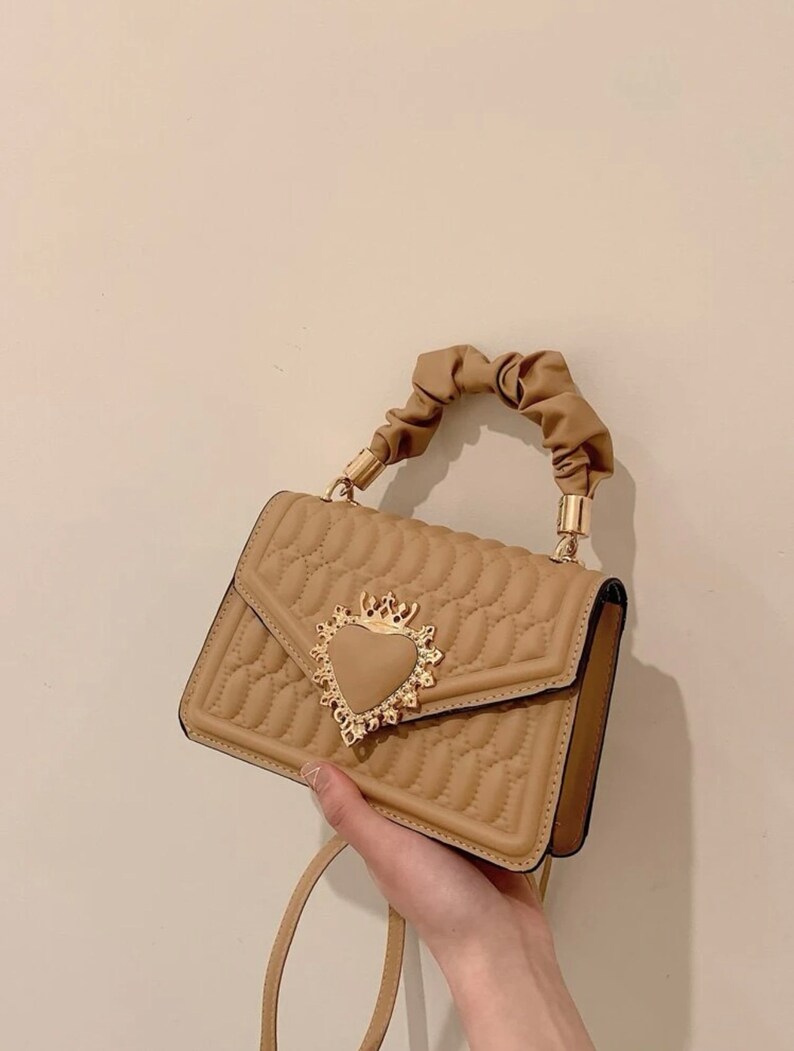 Puede incluir: Un bolso de mano marr&oacute;n acolchado con un cierre dorado en forma de coraz&oacute;n y un asa superior anudada. El bolso tiene una correa larga.