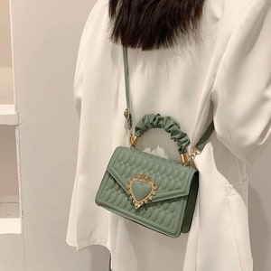 Puede incluir: Un peque&ntilde;o bolso de mano verde acolchado con un cierre de coraz&oacute;n dorado y un asa superior. El bolso tiene una correa larga para llevarlo al hombro.