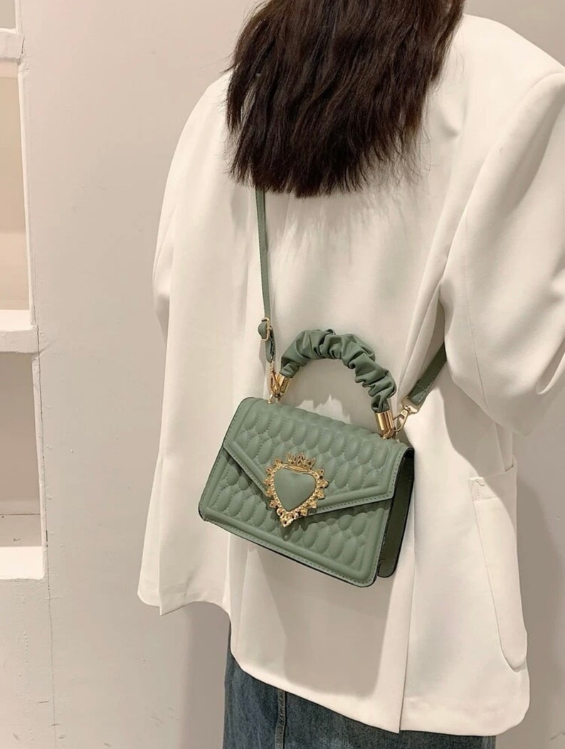 Puede incluir: Un peque&ntilde;o bolso de mano verde acolchado con un cierre de coraz&oacute;n dorado y un asa superior. El bolso tiene una correa larga para llevarlo al hombro.