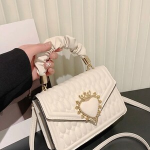 Puede incluir: Un bolso blanco acolchado con un cierre en forma de coraz&oacute;n y un asa con volantes. El cierre tiene una corona y pedrer&iacute;a.