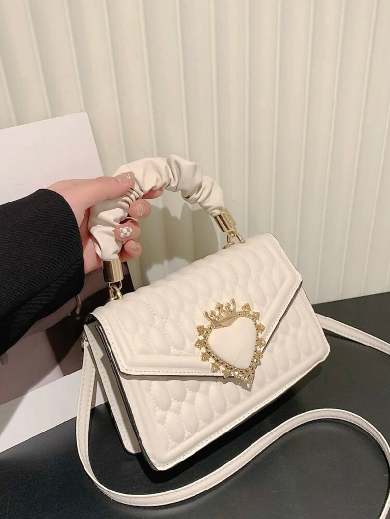 Puede incluir: Un bolso blanco acolchado con un cierre en forma de coraz&oacute;n y un asa con volantes. El cierre tiene una corona y pedrer&iacute;a.