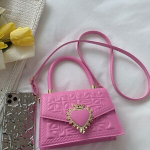 Puede incluir: Un bolso de mano acolchado de color rosa con un cierre en forma de coraz&oacute;n dorado y una correa de cadena. El bolso tiene un asa superior y una correa de hombro desmontable.