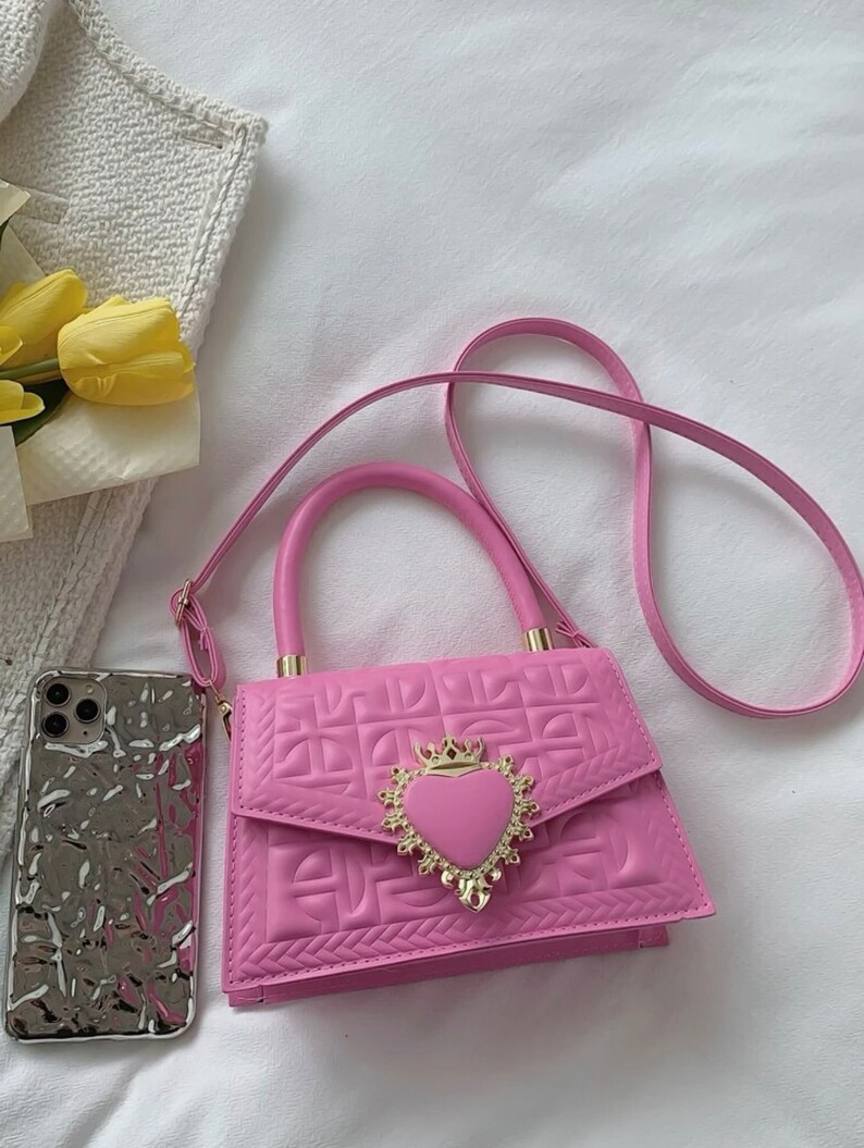 Puede incluir: Un bolso de mano acolchado de color rosa con un cierre en forma de coraz&oacute;n dorado y una correa de cadena. El bolso tiene un asa superior y una correa de hombro desmontable.