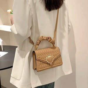 Puede incluir: Un bolso de mano acolchado de color beige con un cierre dorado en forma de coraz&oacute;n y un asa de cuerda trenzada. El bolso tiene una correa larga para llevarlo al hombro.
